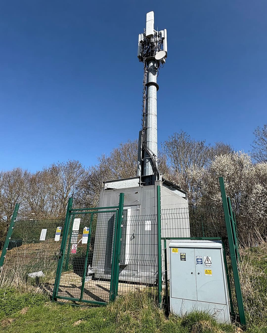 Mastro de antenas de telefonia celular cercado por grade, com armários de equipamentos na base, numa área rural